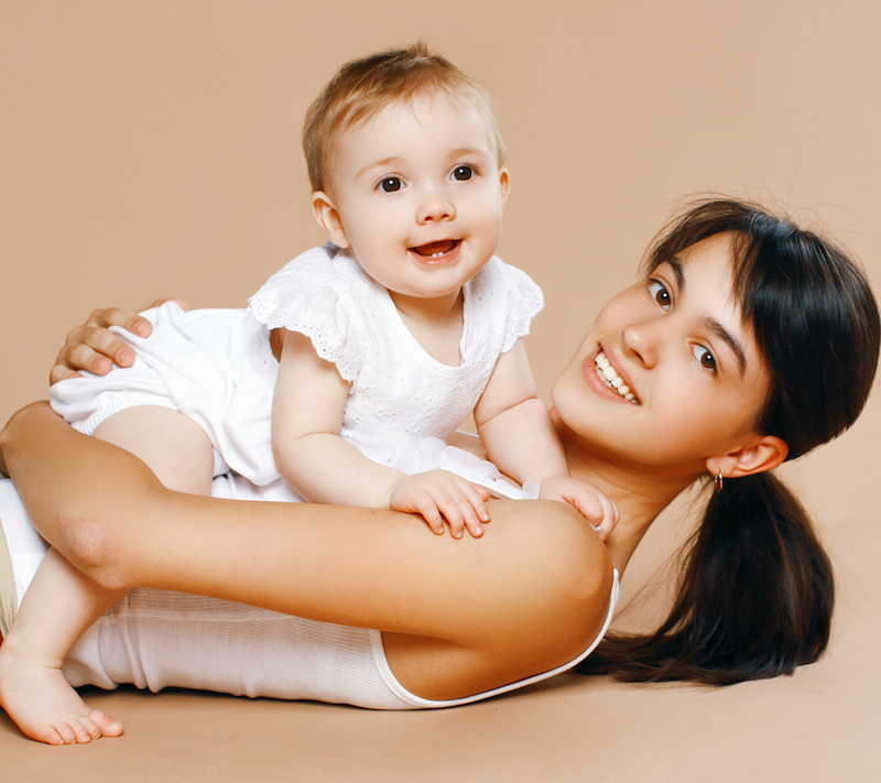 Mother holding smiling baby in arms while laying down looking towards you.